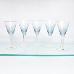 CRISTAL HERING -BLUMENAU. Elegante conjunto de taças para água em cristal, fabricação nacional, circa 1960-70. Bojo de formato obcônico com transição cromática na tonalidade azul-baikal translúcida na porção inferior, terço superior exibe lapidação em sulcos verticais paralelos, fuste hexagonal sobre base circular. Composto de cinco(05) unidades. Dois exemplares conservam as etiquetas de manufatura originais. Medidas: Alt.: 17,5 x Diam.: 9 cm.