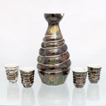 Conjunto para saquê em porcelana moldada, composto por uma(01) garrafa(tokkuri) e quatro(04) copos(choko), de procedência oriental, meados do século XX. Apresentam corpo com conformação anelada helicoidal em relevo recoberta por vidrado saturado de ferro com iridescência grafite, interceptado por filetes dourados brunidos nas arestas e decoração pictórica policromada de folhas de momiji(bordo japonês) em tons de verde, amarelo e terracota. O interior dos recipientes exibe esmaltação branca. Garrafa apresenta quebradura na borda, e copos com desgaste da esmaltação, vide foto. Medidas: Garrafa: Alt.: x Diam.: cm; Copo: Alt.: x Diam.: cm.