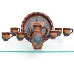 Serviço para café em cerâmica policromada, produção artesanal. Conjunto composto por oito(08) peças: uma cafeteira com tampa, seis xícaras e uma bandeja circular. Modelagem em cerâmica de tonalidade terracota, com decoração incisa e pintada em azul acinzentado, alaranjado, creme e castanho-escuro, formando composições florais e geométricas distribuídas pelos corpos das peças. Cafeteira de formato alongado, com bico curvo, alça lateral e tampa rematada por pega cônica. Bandeja circular com aba elevada e ornamentação correspondente. Xícaras de corpo bojudo, base anelar e alças curvas, repetindo o padrão decorativo do conjunto. Medidas bule: Alt.: 30 x Comp.: 19,5 x Larg.: 11,5cm; Medidas caneca: Alt.: 8 x Comp.: 11 x Larg.: 8 cm; Medida bandeja: Diam.: 35,5 cm.