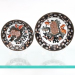 Dois(02) pratos decorativos de parede em porcelana chinesa com guarnição em latão, estilo Imari, circa meados do século XX. Cada peça exibe centro ornamentado com arranjo floral de crisântemos e peônias em policromia sob o vidrado, utilizando paleta em tons de vermelho-ferro, preto e cinza. Aba circular delimitada por cercadura perimetral escura contendo flores brancas e folhagens estilizadas. O reverso apresenta montagem estrutural em liga de latão que recobre as bordas e o bojo externo, contendo argola integrada para suspensão em parede. O fundo da porcelana ostenta marca circular carimbada em tom vermelho com monograma central "CD" envolto pela inscrição "Made in China / For Decorative Use". Medidas: Diam.: 15 cm; Diam.: 12,5cm.