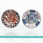 Dois(02) pratos de parede decorativos em porcelana chinesa com guarnição em latão, estilos Imari e Família Rosa, circa meados do século XX. O primeiro exemplar exibe composição floral central com crisântemos em vermelho-ferro e cercadura perimetral escura. O segundo ostenta ao centro a representação zoomórfica de um dragão imperial em tons de azul e vermelho entre nuvens estilizadas, ladeado na aba por reservas com fênix (fenghuang) em tom rosa e painéis geométricos em azul sob o vidrado. Ambos apresentam montagem estrutural externa em latão dotada de argola para suspensão e fundo com marca circular carimbada contendo o monograma "CD" e a inscrição "Made in China / For Decorative Use". Medidas: Diam.: 10cm.