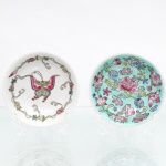 Dois(02) pratos de parede decorativos em porcelana chinesa, estilo Família Rosa e Munshou, circa meados do século XX. O primeiro exemplar de fundo branco abriga representação zoomórfica central de uma borboleta (hudie) com asas expandidas em esmalte rosa e verde, circundada por grinalda perimetral de ramificações fitomórficas e botões florais interligados por fitas. O segundo apresenta fundo esmaltado na tonalidade verde-turquesa, densamente decorado por peônias, crisântemos e flores diversas dispersas em arranjo livre. O reverso de ambas as peças exibe base anelar em desprovida de esmalte e fixação artesanal de passador metálico(adaptado) triangular por meio de massa adesiva cinza(durepox) para suspensão em parede, obliterando parcialmente possíveis marcas de fábrica subjacentes. Medidas: Diam.: 10cm.
