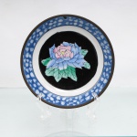 Prato de parede decorativo em porcelana chinesa com guarnição em latão, estilo Cantão de fundo preto (Famille Noire), circa meados do século XX. O centro abriga uma grande peônia esmaltada em tons de azul, rosa, amarelo e verde sobre um medalhão circular de fundo negro opaco, simbolizando riqueza e honra. A aba larga exibe decoração geométrica texturizada em azul e branco sob o vidrado, emulando uma padronagem de mosaico ou gelo fendido(cracked ice). O reverso apresenta montagem estrutural externa em liga de latão dotada de passador rebitado para suspensão em parede, além de fundo com marca de manufatura circular carimbada e a inscrição "Made in China / For Decorative Use". Medidas: Alt.: 3,5 x Diam.: 15 cm.