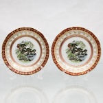 Porcelana, Steatita e Magnésia S/A, Campo Largo - PR(Fundada em 1954). Par de pratos decorativos em porcelana nacional, circa 1950-1960. Apresentam formato circular com aba recortada em ondulações, ornamentada por debrum vermelho-vinho e guirlandas florais em ouro. Centro com decoração policromada com paisagem oriental composta por pescador em embarcação, ponte de pedra, pavilhões e montanhas ao fundo. O reverso exibe suportes metálicos para suspensão e marca de manufatura. Medida: Diam.: 15 cm.