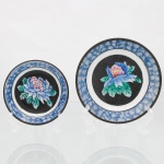 Dois(02) pratos de parede decorativos em porcelana chinesa com guarnição em latão, estilo Cantão de fundo preto (Famille Noire), circa meados do século XX. O centro de cada peça abriga uma grande peônia esmaltada em tons de azul, rosa, amarelo e verde sobre um medalhão circular de fundo negro opaco, simbolizando riqueza e honra. A aba larga exibe decoração geométrica texturizada em azul e branco sob o vidrado, emulando uma padronagem de mosaico ou gelo fendido(cracked ice). O reverso apresenta montagem estrutural externa em liga de latão dotada de passador rebitado para suspensão em parede, além de fundo com marca de manufatura circular carimbada e a inscrição "Made in China / For Decorative Use". Medidas: Diam.:  10cm; Diam.: 13cm.