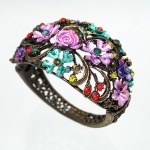 Bracelete rígido executado em metal vazado de tonalidade bronze, com composição formada por volutas, ramagens e elementos florais aplicados em relevo. Flores esmaltadas em tons de rosa, violeta, verde e azul-turquesa, intercaladas por cabochões e pedras de imitação facetadas nas cores vermelha, verde, amarela, azul e incolor. Estrutura articulada por dobradiça lateral, com superfície interna igualmente trabalhada em motivos vegetalistas. Apresenta oxidação e perdas pontuais de esmaltação compatíveis com o uso. Medidas: Alt.: 6,5 x Comp.: 8 x Larg.: 5cm.