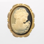 Broche camafeu de figura feminina, composto por camafeu oval em material sintético moldado, de fundo de tonalidade preto dégradé e relevo marfim. Representação de busto feminino de perfil voltado à direita, com penteado preso e ramalhete de rosas junto ao colo. Montagem em moldura de metal dourado, de contorno lobulado e friso perlado. Reverso provido de alfinete basculante com trava. Medida: