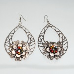 BIJUTERIA. Par de brincos pendentes com ornamentação floral, executados em metal de tonalidade prateada com acabamento oxidado. Estrutura pendente em formato de gota, constituída por trama vazada de motivos geométricos e curvilíneos, sobreposta na porção inferior por composição floral em camadas. Centros guarnecidos por strass facetados em vermelho, âmbar, verde, azul, rosa e incolor. Suspensão por anzóis metálicos para orelha perfurada; reversos revelam construção estampada e montagem por sobreposição. Medidas: Comp.: 8 x Larg.: 5,5cm.