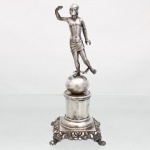 Prata. Porto, terceiro quartel do século XIX. Excepcional paliteiro de coleção confeccionado em prata portuguesa, c.1853 - 1861. Composto por coluna de composição arquitetônica e figurativa. Base quadrangular assente sobre quatro pés fundidos em forma de garra, ornamentados por folhagens, flores e volutas em relevo; corpo central constituído por plinto escalonado e receptáculo cilíndrico facetado, encimado por placa circular perfurada destinada à disposição dos palitos. Sobre esfera se ergue a figura mitológica de Mercúrio(Hermes), executada em vulto pleno, representado com torso descoberto, panejamento cingindo os quadris e pétaso alado sobre a cabeça. Junto aos tornozelos observam-se talárias, sandálias aladas, atributos iconográficos associados ao mensageiro dos deuses. Figura em atitude dinâmica, com um dos braços elevado e o outro descendente. Apresenta notável perda do caduceu, uma asa no pétaso e outra no lado externo do pé direito. Apresenta contraste "P Coroa" e punção de ourives. Medidas: Alt.: x Comp.: x Larg.:; Peso: