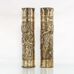 Par de vasos confeccionados a partir de estojos de munição de artilharia em latão, arte de trincheira (trench art). Corpos cilíndricos obtidos pelo reaproveitamento de estojos militares, trabalhados por martelagem e repuxo, com decoração vegetalista em relevo composta por ramos de videira, folhas e cachos de uvas sobre campo inteiramente texturizado. Faixas superiores ornamentadas por motivos foliares estilizados e reservas geométricas, com superfícies alternadamente polidas e marteladas. Bases conservam as marcações originais dos estojos, entre elas "75 DEC, MGM 58, L.17 BX", além de inscrições e marcas junto às espoletas. Trabalho decorativo realizado sobre material militar reutilizado, segundo tradição difundida na chamada trench art. Medidas: Alt.: 35 x Diam.: 8,5 cm.