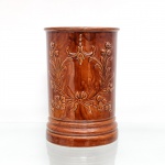 Vaso confeccionado em cerâmica vidrada. Corpo cilíndrico, bordo saliente e base escalonada, revestido por esmalte em tonalidade castanha. Superfície ornamentada em relevo com ramos, flores, folhagens, laços e elementos fitomórficos, distribuídos em composições contínuas ao redor do corpo. Interior esmaltado. Medidas: Alt.: x Diam.: