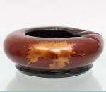 Cinzeiro circular em cerâmica, Corpo baixo de formato anular, revestido por esmalte em tonalidade vermelho-acastanhada, com base e contorno interno em preto. Bordo provido de três reentrâncias destinadas ao apoio de cigarros. Superfície externa ornamentada por aplicações irregulares em tonalidade dourada. Base sem marca identificada. Medidas: Alt.: x Diam.: