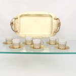 Serviço para café em porcelana e metal dourado. Composto por seis(06) xícaras cilíndricas em porcelana branca, montadas em suportes metálicos vazados em padrão reticulado e providos de alça lateral, seis pires circulares com decoração floral em relevo, e uma(01) bandeja retangular de cantos arredondados, centro ligeiramente rebaixado, com duas alças laterais modeladas em cordão e elementos foliares. Acabamento metálico em tonalidade dourada. Estado de conservação conforme fotografias. Medidas: