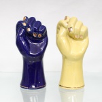 Duas fígas executadas em cerâmica esmaltada, moldadas em vulto pleno, uma sob esmalte azul-cobalto e outra em amarelo-claro. Unhas realçadas em dourado e filete da mesma tonalidade junto às bases. Corpos ocos, com abertura inferior e pequeno orifício posterior. Medidas: Alt.: 17,5 x Comp.: 8,5 x Larg.: 7cm.