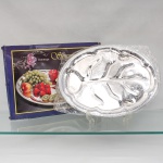 Travessa oval para servir em metal prateado, acompanhada de caixa original, possui borda trabalhada em friso estriado e centro compartimentado em formato estilizado de espinha de peixe com canais de escoamento e gravação fitomórfica em baixo-relevo. Acondicionado em emabalagem e caixa original, sem uso. Medidas: Comp.: 45,5 x Larg.: 34cm.