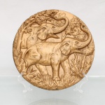 Prato decorativo de parede em chapa metálica conformada em alto-relevo, com acabamento em pátina dourada. A peça apresenta composição iconográfica representando dois elefantes com as trombas erguidas em cenário de floresta tropical. O reverso exibe o contra-relevo negativo resultante do processo de prensagem, provido de argola de suspensão soldada. Medida: Diam.: 22 cm.