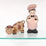 Dois(02) porta-utensílios de cozinha em cerâmica policromada, modeladas em formas figurativas de cozinheiro e vaca, com corpos ocos e aberturas destinadas à acomodação de colheres e demais utensílios culinários. Figura do cozinheiro representada com toque, avental, gravata-borboleta e sapatos, em tonalidades rosa, cinza e preto; figura bovina com pintura em tons de marrom, detalhes faciais policromados. Execução moldada e pintura manual.Medidas: Alt.: 25,5 x Comp.: 12 x Larg.: 9cm.Medidas: Alt.: 11 x Comp.: 12 x Larg.: 12,5cm.