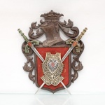 Panóplia heráldica decorativa em resina e metal, estilo revivalista, arranjo estruturado sobre placa de suporte moldada exibindo elmo com timbre coroado ladeado por paquifes em relevo simétrico, centro ostenta escudo heráldico recortado sobre fundo têxtil vermelho, contendo brasão de armas com águia bicéfala e escudete quadrifoliado esmaltado. Duas espadas cruzadas em sautor complementam o conjunto, apresentando lâminas com gravuras lineares em baixo-relevo, guardas arqueadas e pomos com formato estilizado de elmo, exibindo policromia e pátina imitativa de oxidação e pátina de bronze antiga. Reverso apresenta fechamento em placa plana com fixação por parafusos centrais e argola de suspensão.Medidas: Comp.: 35,5 x Larg.: 28 cm. Estado conforme fotografias, apresenta sinais de uso e do tempo.