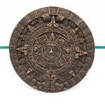 Placa decorativa circular em resina moldada, reproduzindo a iconografia da Pedra do Sol (Calendário Asteca), circa segunda metade do século XX. Centro ostenta a efígie em relevo pronunciado do deus Tonatiuh com a língua glotografada em formato de faca de sacrifício (tecpatl), circundado por anéis concêntricos que delimitam os quatro glifos das eras precedentes(Olin) e os vinte dias do ciclo tonalpopohualli, periferia exibe raios solares alternados com representações simbólicas e duas serpentes de fogo(Xiuhcoatl) contrapostas na base inferior; superfície apresenta pátina escura imitativa de rocha basáltica com incrustações terrosas artificiais. Verso plano possui revestimento em tecido marrom dotado de passador metálico para fixação em parede. Medidas: Alt.: 3 x Diam.: 29,5 cm.