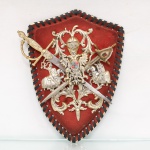 Panóplia heráldica decorativa em metal e madeira, de fabricação portuguesa, arranjo estruturado sobre placa de formato de escudo recortado apresenta revestimento frontal em veludo vermelho com borda em tira de couro trançada, ornamentação central em metal dourado e prateado exibe elmo com visor de grades encimado por paquifes fitomórficos, assentado sobre águia bicéfala heráldica; peito da ave ostenta escudete esquartelado esmaltado nas tonalidades azul e vermelha, conjunto ladeado por duas figuras em relevo de cavaleiros medievais justadores e cortado em sautor por duas espadas, cujas lâminas de aço trazem a inscrição "Portugal" em cartela cinzelada. Verso em placa de fibra de madeira exibe orifício superior para fixação em parede. Estado de conservação conforme fotografias. Medidas: Comp.: 30 x Larg.: 22 cm.
