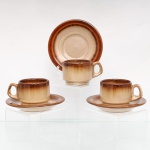 Três(03) xícaras para café em cerâmica de grés de alta resistência, manufaturados pela oficina Oxford, fabricação nacional, c.1970-80. Bojo cilíndrico e alça lateral estilizada, apresentam esmaltagem vítrea acetinada com fundo em tom areia salpicado por minúsculas pontuações escuras, resultado da queima de minerais de ferro contidos na própria argila. As bordas da xícara e do pires recebem decoração perimetral degradê em esmalte escorrido na cor castanha, resultando em transições lineares e nuances concêntricas diferenciadas para cada exemplar. O verso exibe base com anel de assento aparente e carimbo de manufatura na cor azul sob o vidrado com as inscrições: "Grés / Oxford / Made in Brazil / 4830". Medidas: Xícara: Alt.: 4,5 x Comp.: 8,5 x Larg.: 6 cm; Pires: Diam.: 11,5 cm.