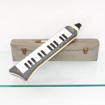 Melódica Hohner, modelo Melodica Piano 26, Alemanha, circa 1950-1960. Instrumento aerofone de palhetas livres, acionado por sopro através de bocal e teclado de 26 teclas, abrangendo pouco mais de duas oitavas. Corpo em material sintético nas tonalidades cinza e marfim, teclado cromático e correia posterior para sustentação durante a execução. Identificação frontal "HOHNER melodica piano 26" e marca da manufatura no reverso. Acompanha estojo de transporte com ferragens metálicas. Apresenta perdas localizadas do revestimento superficial do corpo. O modelo integra a primeira geração de melódicas desenvolvidas e difundidas pela Hohner em meados do século XX; exemplar correspondente conservado no National Museum of American History, Smithsonian Institution, é catalogado como produção alemã de circa 1950-1960. No estado. vide fotos. Medidas: Alt.: 41,5 x Comp.: 9 x Larg.: 4,5cm.