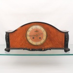 Westerstrand, Töreboda - Suécia. (Fundada em 1906). Relógio de mesa, fabricação sueca, período entre-guerras, circa década de 1930. A caixa de perfil arqueado apresenta acabamento folheado em madeira com veios longitudinais aparentes, delimitada por debrum superior arqueado e saia inferior recortada com motivos fitomórficos em madeira ebonizada.  As laterais ostentam montantes aplicados com entalhes vazados em relevo folhoso de acanto negro. O mostrador circular, protegido por aro de latão e vidro bombée articulado, traz numeração arábica sobre coroa marfim, centro folheado a madeira, ponteiros vazados e plaqueta metálica trapezoidal com a marca do fabricante. O reverso abriga portinhola que resguarda maquinário em latão com sistema de badalo por martelo sobre gongo e pêndulo suspenso, contendo punção circular na platina traseira. A estrutura denota perdas significativas de madeira e folheado na aresta superior esquerda da curvatura, oxidação nos componentes metálicos e marcas de atrito. Falta pêndulo. Apresenta avarias na estrutura da caixa, vide foto. Medidas: Alt.: 25 x Comp.: 55,5 x Larg.: 16,5 cm.
