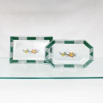Conjunto de duas bandejas em vidro prensado e decorado, formato retangular e outro oitavado, ambos de centro rebaixado com textura reticulada. Abas ornamentadas por faixa geométrica em verde, alternando campos lisos e segmentos lineares. Ao centro, composição floral em estampagem policromada com flores, botões, folhas e hastes em amarelo, vermelho, azul e verde. Medidas: Comp.: x Larg.: cm; Comp.: x Larg.: cm.
