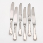 MERIDIONAL EPNS 100. Elegante conjunto de facas médias confeccionadas em metal espessurado à prata(EPNS 100), cabo violinado com filetes longitudinais em baixo-relevo e terminação ornamentada por composição de concheados, volutas, flores e folhagens em relevo. Lâmina em aço inox. Marca da manufatura gravada na lâmina, e identificação EPNS 100 puncionada no cabo. Composto de seis(06) unidades. Medida: Comp.: 21 cm.
