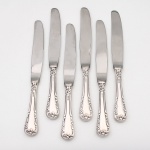 MERIDIONAL EPNS 100. Elegante conjunto de facas médias confeccionadas em metal espessurado à prata(EPNS 100), cabo violinado com filetes longitudinais em baixo-relevo e terminação ornamentada por composição de concheados, volutas, flores e folhagens em relevo. Lâmina em aço inox. Marca da manufatura gravada na lâmina, e identificação EPNS 100 puncionada no cabo. Composto de seis(06) unidades. Medida: Comp.: 21 cm.