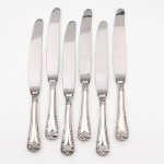 MERIDIONAL EPNS 100. Elegante conjunto de facas grandes confeccionadas em metal espessurado à prata(EPNS 100), cabo violinado com filetes longitudinais em baixo-relevo e terminação ornamentada por composição de concheados, volutas, flores e folhagens em relevo. Lâmina em aço inox. Marca da manufatura gravada na lâmina, e identificação EPNS 100 puncionada no cabo. Composto de seis(06) unidades. Medida: Comp.: 24 cm.
