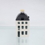 Garrafa miniatura em porcelana moldada em forma de casa holandesa, pertencente à série de casas Delft produzidas para a KLM (Koninklijke Luchtvaart Maatschappij, companhia aérea neerlandesa fundada em 1919) e destinada ao acondicionamento de Bols Genever(destilado tradicional neerlandês à base de grãos e aromatizado com zimbro, produzido pela casa Lucas Bols, fundada em Amsterdã em 1575). Corpo arquitetônico de planta retangular, com fachadas moldadas em relevo simulando alvenaria, portas e janelas, cobertura inclinada com telhas em relevo e chaminé utilizada como gargalo, fechada por tampa. Esmaltação vítrea em branco, azul-escuro e azul acinzentado. Verso com as inscrições 55, Bols Amsterdam 1575 e logotipo KLM. Base com inscrição Blue Delfts exclusively made for KLM by Bols Amsterdam 1575  Bols Genever 35 ml e numeração moldada 2022 55. KLM House nº 55. Medidas: Alt.: 11 x Comp.: 6,5 x Larg.: 5,5cm.