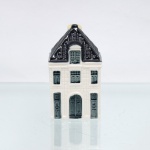 Garrafa miniatura em porcelana moldada em forma de casa holandesa, pertencente à série de casas Delft produzidas para a KLM (Koninklijke Luchtvaart Maatschappij, companhia aérea neerlandesa fundada em 1919) e destinada ao acondicionamento de Bols Genever(destilado tradicional neerlandês à base de grãos e aromatizado com zimbro, produzido pela casa Lucas Bols, fundada em Amsterdã em 1575). Corpo arquitetônico de planta retangular, com fachadas moldadas em relevo simulando alvenaria, portas e janelas, cobertura inclinada com telhas em relevo, águas-furtadas e chaminé utilizada como gargalo, fechada por tampa. Esmaltação vítrea em branco, azul-escuro e azul acinzentado. Verso com as inscrições 64, Bols Amsterdam 1575 e logotipo KLM. Base com inscrição Blue Delfts exclusively made for KLM by Bols Amsterdam 1575  Bols Genever 35 ml e numeração moldada 2022 64. KLM House nº 64. Medidas: Alt.: 10 x Comp.: 6 x Larg.: 5,5cm.