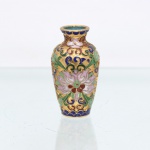 Vaso miniatura decorativo em metal esmaltado com a técnica de cloisonné. Corpo em formato balaústre assentado sobre base circular plana, recoberto por fundo em ouro fosco obtido por fios de latão alveolados (cloisons). A superfície apresenta ornamentação contínua de caráter floral e fitomórfico, destacando grande flor de lótus central estilizada com pétalas nas tonalidades lilás e branca, acompanhada por ramagens simétricas em esmalte verde, detalhes espiralados em azul-cobalto e pequenas corolas em vermelho-óxido. O colo e a base são arrematados por cercaduras geométricas semicirculares simulando escamas em tom azul. O interior do bocal e o verso da base exibem contra-esmaltagem lisa em tom azul-turquesa uniforme. Medidas: Alt.: 7,5 x Diam.: 4cm.