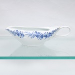 Real  Indústria Brasileira de Porcelanas, Mauá  SP (fundada em 1943 por Fritz Erwin Schmidt). Molheira em porcelana refratária, modelo Calorama Super Refratária, de corpo alongado, bojo profundo, bico vertedor afilado e alça lateral curvada. Esmaltação vítrea na cor branca, com decoração fitomórfica em azul, composta por frisos de flores, folhas e ramos distribuídos longitudinalmente nas duas faces. Base com marca de fábrica sob coroa e inscrições. Medidas: Alt.: 7 x Comp.: 25,5 x Larg.: 11cm.