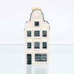 Garrafa miniatura em porcelana moldada em forma de casa holandesa, pertencente à série de casas Delft produzidas para a KLM (Koninklijke Luchtvaart Maatschappij, companhia aérea neerlandesa fundada em 1919) e destinada ao acondicionamento de Bols Genever(destilado tradicional neerlandês à base de grãos e aromatizado com zimbro, produzido pela casa Lucas Bols, fundada em Amsterdã em 1575). Corpo arquitetônico de planta retangular, com fachadas moldadas em relevo simulando alvenaria, portas e janelas, cobertura inclinada com telhas em relevo e chaminé utilizada como gargalo, fechada por tampa. Esmaltação vítrea em branco, azul-escuro e azul acinzentado. Verso com a númeração 72 sobre as inscrições Bols Amsterdam 1575 e logotipo KLM. Base com inscrição "Blue Delfts exclusively made for KLM by Bols Amsterdam 1575 ", "HKDNP" e numeração em baixo-relevo "2012 - 74". Medidas: Alt.: 9,5 x Comp.: 4 x Larg.: 5cm. (Esta 74ª casa da série KLM está localizada no número 87 da Singel , em Amsterdã, construída por volta de 1730).