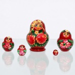 Conjunto de bonecas tradicionais russas do tipo matryoshka, confeccionado em madeira torneada e pintada à mão. Composto por cinco figuras encaixáveis de dimensões decrescentes, o grupo apresenta acabamento em verniz brilhante sobre fundo de tonalidade vermelha uniforme. A decoração exibe feições femininas juvenis com bochechas coradas, olhos amendoados e cabelos castanhos claros sob lenço de cabeça (shashura) adornado por pequenas flores brancas de miolo amarelo e ramagens simplificadas. O ventre das figuras destaca arranjo floral proeminente composto por grandes rosas em tons de rosa e vermelho com detalhes em ouro fosco, ladeadas por densas folhas verdes em pinceladas livres. Medidas: Alt.: 8 x Diam.: 5cm.Medidas: Alt.: 5,5 x Diam.: 4cm.Medidas: Alt.: 3,5 x Diam.: 3cm.Medidas: Alt.: 2 x Diam.: 2cm.Medidas: Alt.: 1 x Diam.: 1cm.