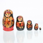 Conjunto de bonecas tradicionais russas do tipo matryoshka, confeccionado em madeira torneada e pintada à mão. Composto por cinco figuras encaixáveis de dimensões decrescentes, o grupo apresenta acabamento em verniz brilhante sobre fundo dividido entre a cor preta no tronco e vermelho translúcido na base, delimitada por um contorno lobulado escuro no reverso. A decoração exibe feições femininas juvenis com bochechas pontuadas, olhos destacados e cabelos avermelhados sob lenço de cabeça (shashura) recoberto por folhas douradas estilizadas e contornos pretos. O ventre das figuras é preenchido por densa composição pictórica de inspiração Khokhloma, contendo arranjo de flores em tons alaranjados e dourados ladeadas por folhagens escuras e pequenas bagas circulares. O reverso do lenço na peça principal repete o padrão floral folheado em ouro fosco. Medidas: Alt.: 8,5 x Diam.: 4,5cm.Medidas: Alt.: 6 x Diam.: 3cm.Medidas: Alt.: 4 x Diam.: 2cm.Medidas: Alt.: 2,5 x Diam.: 1,5cm.Medidas: Alt.: 1 x Diam.: 0,5cm.