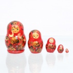 Conjunto de bonecas tradicionais russas do tipo matryoshka, confeccionado em madeira torneada e pintada à mão. Composto por cinco figuras encaixáveis de dimensões decrescentes, o grupo apresenta acabamento em verniz sobre fundo de tonalidade vermelha uniforme. A decoração exibe feições femininas juvenis com bochechas pontuadas, olhos destacados e cabelos castanhos claros sob lenço de cabeça (shashura) decorado por folhas estilizadas em tons dourados e marrons com contornos pretos. O ventre das figuras é preenchido por composição pictórica tradicional de inspiração Khokhloma, contendo arranjo de flores douradas circundadas por folhagens escuras e uma seção inferior em padrão de treliça geométrica preta entrelaçada. O reverso da saia na peça principal apresenta uma cercadura de arcos festonados em tons claros e dourados. Medidas: Alt.: 7,5 x Diam.: 5cm.Medidas: Alt.: 5,5 x Diam.: 3,5cm.Medidas: Alt.: 3,5 x Diam.: 2,5cm.Medidas: Alt.: 2 x Diam.: 1,5cm.Medidas: Alt.: 1 x Diam.: 0,5cm.
