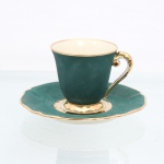 Xícara para café em porcelana francesa de Limoges, manufaturada pela tradicional oficina de R. Leclair (Leclair Porcelain), datável do período compreendido entre 1949 e 1954. Bojo campaniforme com bocal expandido e pires de aba recortada. A superfície estrutural externa da xícara e o espelho superior do pires recebem cobertura homogênea de esmalte fosco em tom verde-esmeralda, simulando textura acetinada. A alça lateral em formato de voluta arqueada, o pé pedestal anelar baixo e o debrum periférico de ambas as peças recebem filetagem à ouro, padrão decorativo que se repete no anel central de contenção do pires. O interior da xícara exibe acabamento em esmalte brilhante de tom marfim. Verso apresenta base circular esmaltada ostentando o sinete de manufatura transferido em pigmento verde, composto por raios solares divergentes sobre as inscrições L E C / IMOGES / France, com a letra inicial "L" estendida de forma estilizada sob o nome da localidade. Medidas: Alt.: 6,5 x Comp.: 8,5 x Larg.: 7cm; Pires: Diam.: 13,5cm.