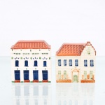 Par de esculturas miniaturizadas em faiança representando edifícios históricos de Amsterdã, manufaturadas pela oficina Elksva, na Holanda, durante a segunda metade do século XX. As estruturas cerâmicas apresentam esmalte branco-marfim com craquelê natural e telhados facetados em marrom-alaranjado. O primeiro exemplar exibe fachada neoclássica com janelas em azul-cobalto e ornamentação fitomórfica verde; o segundo apresenta frontão escalonado barroco, portal em arco pleno e janelas azuladas. O verso de ambos exibe carimbo azul da manufatura com coroa heráldica, a inscrição "Delftware Handwerk Designed by Elksva Holland"" e os códigos de série "BS-4" e "BS-6". Medidas: Alt.: 6,5 x Comp.: 6 x Larg.: 3cm.