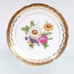 Porcelana Real, Mauá - SP(Fundada em 1943 por Fritz Erwin Schmidt). Prato raso em porcelana nacional, circa 1960-1970. Esmaltagem vítrea na cor branca, aba recortada em ondulações, ornamentada por larga faixa vermelho-bordeaux com reservas losangulares e florões em ouro, seguidos por uma cercadura interna festonada de pequenas ramagens e gavinhas douradas. Centro abriga arranjo pictórico policromado por processo de decalcomania, representando buquê assimétrico com rosas, crisântemos, violetas e folhagens detalhadas, delimitado por um fino filete circular metalizado. Marca da manufatura no verso. Marca da manufatura no verso. Medida: Diam.: 23 cm.