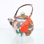 Bule em cerâmica japonesa Banko, com ornamentação de fauna marinha em alto-relevo. Japão, final do período Meiji/início do período Taisho, circa 1900-1920. Recipiente de conformação globular, modelado e esmaltado em policromia, revestido por composição marinha constituída por peixes, crustáceos e moluscos aplicados em relevo, entre os quais se distinguem lagosta, caranguejo, diferentes espécies de peixes e concha, esta última conformando o bico vertedor. Tampa modelada como peixe achatado, com olhos destacados e pomo em forma de fruto. Alça superior arqueada em junco ou fibra vegetal, presa ao corpo por passadores cerâmicos. Superfícies trabalhadas com incisões, pontilhados e relevos destinados à caracterização de escamas, carapaças e demais elementos anatômicos, sob esmaltes em vermelho, verde, azul, rosa, cinza, castanho e amarelo. Produção japonesa de caráter naturalista associada às cerâmicas Banko destinadas ao mercado de exportação, circa início do século XX. Apresenta perdas e desgastes compatíveis com o período, observáveis na tampa, elementos aplicados e bordas. Medidas: Alt.: 16,5 x Comp.: 14,5 x Larg.: 11cm.