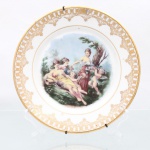 Porcelana Real, Mauá - SP(Fundada em 1943 por Fritz Erwin Schmidt). Prato decorativo de parede em porcelana naciona, circa 1960-1970). Esmlatagem vítrea na cor branca, aba exibe cercadura em ouro  constituída por friso geométrico treliçado e arcadas ogivais, complementada por pingentes inferiores em arabescos festonados e volutas. Centro abriga composição pictórica policromada por processo de decalcomania de inspiração neoclássica e mitológica, retratando três figuras femininas em trajes diáfanos ladeadas por dois cupidos alados(putti) portando guirlandas e cestas de flores em cenário bucólico arborizado. Verso com garra metálica para suspensão e marca de manufatura. Medida: Diam.: 22 cm.