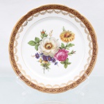 Porcelana Real, Mauá - SP(Fundada em 1943 por Fritz Erwin Schmidt). Prato raso em porcelana nacional, circa 1960-1970. Esmaltagem vítrea na cor branca, aba recortada em ondulações, ornamentada por larga faixa vermelho-bordeaux com reservas losangulares e florões em ouro, seguidos por uma cercadura interna festonada de pequenas ramagens e gavinhas douradas. Centro abriga arranjo pictórico policromado por processo de decalcomania, representando buquê assimétrico com rosas, crisântemos, violetas e folhagens detalhadas, delimitado por um fino filete circular metalizado. Marca da manufatura no verso. Marca da manufatura no verso. Medida: Diam.: 23 cm.