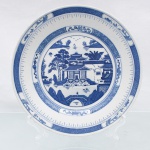 Prato decorativo em porcelana de pasta dura, produzido pela manufatura Renmin Cichang (Fábrica de Porcelana do Povo) em Jingdezhen, China, durante a segunda metade do século XX, circa 1969-1990. Esmaltagem vítrea na cor branca, apresentando aba perimetral recoberta por decorações transferidas sob o vidrado em tom azul-cobalto. A ornamentação exibe o célebre padrão "Blue Willow" de inspiração oriental Chinoiserie, guarnecido por cercaduras concêntricas em padrões de reticulados geométricos, pontilhados lineares e reservas com motivos fitomórficos abstratos instalados ao longo da borda. Centro abriga composição pictórica que reproduz uma gravação, retratando cenário paisagístico composto por pagode tradicionalizado de múltiplos tetos, pontes arqueadas com personagens, embarcações navegando ao fundo, salgueiro árvores frutíferas e o pássaros em revoada na porção superior central. Verso exibe base com anel de assentamento aparente e carimbo de manufatura transferido na cor azul, contendo as inscrições em caracteres chineses (Renmin Cichang) dispostos em arco sobre a localidade produtora (Jingdezhen). Medida: Diam.: 25 cm.