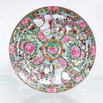 Prato de coleção em porcelana chinesa, seguindo estritamente a gramática ornamental do estilo Medalhão de Cantão da Família Rosa. Superfície interna apresenta organização radial compartimentada a partir de uma reserva central circular circundada por filete dourado, contendo peônia fênix ladeada por ave canora policromada. Da periferia deste núcleo divergem faixas sinuosas que delimitam quatro painéis principais em formato de leque, intermeados por borboletas heráldicas, gavinhas folheadas e botões florais sobrepostos. Duas das reservas opostas abrigam representações minuciosas de cenas palacianas, evidenciando figuras da nobreza em conversação no interior de pavilhões arquitetônicos tradicionais; os dois quadrantes alternados exibem densa composição de peônias abertas, frutos e aves exóticas em ramagens. O reverso apresenta anel de assento plano desprovido de esmalte e sinete APÓCRIFO quadrangular aplicado em pigmento vermelho-ferro sob a inscrição(Qianlong Nian Zhi), alusiva ao reinado da Dinastia Qing. Medida: Diam.: 20,5 cm.
