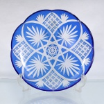 Prato decorativo em cristal lapidado e revestido (overlay), manufaturado na região da Boêmia (atual República Tcheca) durante a segunda metade do século XX. A peça apresenta formato circular com borda superior sinuosa recortada em gomos ondulares, executada em cristal translúcido recoberto por camada externa homogênea em tom azul-cobalto. A ornamentação é obtida por processo de lapidação manual facetada em baixo-relevo que remove a película colorida superficial, revelando a transparência do cristal base. O espelho central abriga um grande medalhão composto por estrela de dez pontas entrelaçada em padrão geométrico concêntrico, circundada por estrutura estelar de linhas cruzadas. A aba e o caveto recebem decoração geométrica simétrica intercalada, composta por quatro cartelas ovais preenchidas por lapidação em bicos de diamante formando reticulados pontiagudos, ladeadas por ramagens fitomórficas estilizadas simulando palmetas e folhas lanceoladas que convergem para a orla. Medidas: Alt.: 4,5 x Diam.: 28,5 cm.