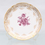 Prato de coleção em porcelana de pasta dura, manufaturado na Europa Ocidental durante o século XIX. Apresenta formato circular com aba ligeiramente recortada em gomos e debrum realçado por filete em ouro, campo central exibe decoração pictórica do tipo "Deutsche Blumen" executada em tonalidade púrpura, representando buquê assimétrico com arranjo de rosas, crisântemos e folhagens detalhadas, aba ostenta rica cercadura ornamental sobreposta em ouro, composta por volutas rococó, ramagens entrelaçadas, cestos suspensos e frisos rendados em guilhoche. O reverso exibe base com anel de assento aparente e medalhão central pintado com botão de rosa em idêntica paleta cromática, ocultando marca de manufatura sob o esmalte. Medida: Diam.: 27 cm.