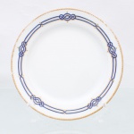 Prato de coleção em porcelana de pasta dura, manufaturado na Europa Ocidental durante o final do século XIX ou início do século XX. Apresenta formato circular plano com aba lisa finalizada em debrum dourado de padrão semicircular repetitivo, aba dotada de ornamentação composta por friso duplo concêntrico em esmalte azul-cobalto sob a técnica de filetagem, intercalado por quatro medalhões geométricos com entrelaçamentos decorativos em nós e realces em ouro, campo central inteiramente liso, ressaltando a brancura da massa vítrea, reverso exibe base com anel de assento concêntrico aparente e numeração "104" pintada à mão livre em pigmento dourado, indicativa de numeração de série, pintor ou padrão fabril.Medida : Diam.: 19cm. Apresenta um ínfimo bicado no bordo.